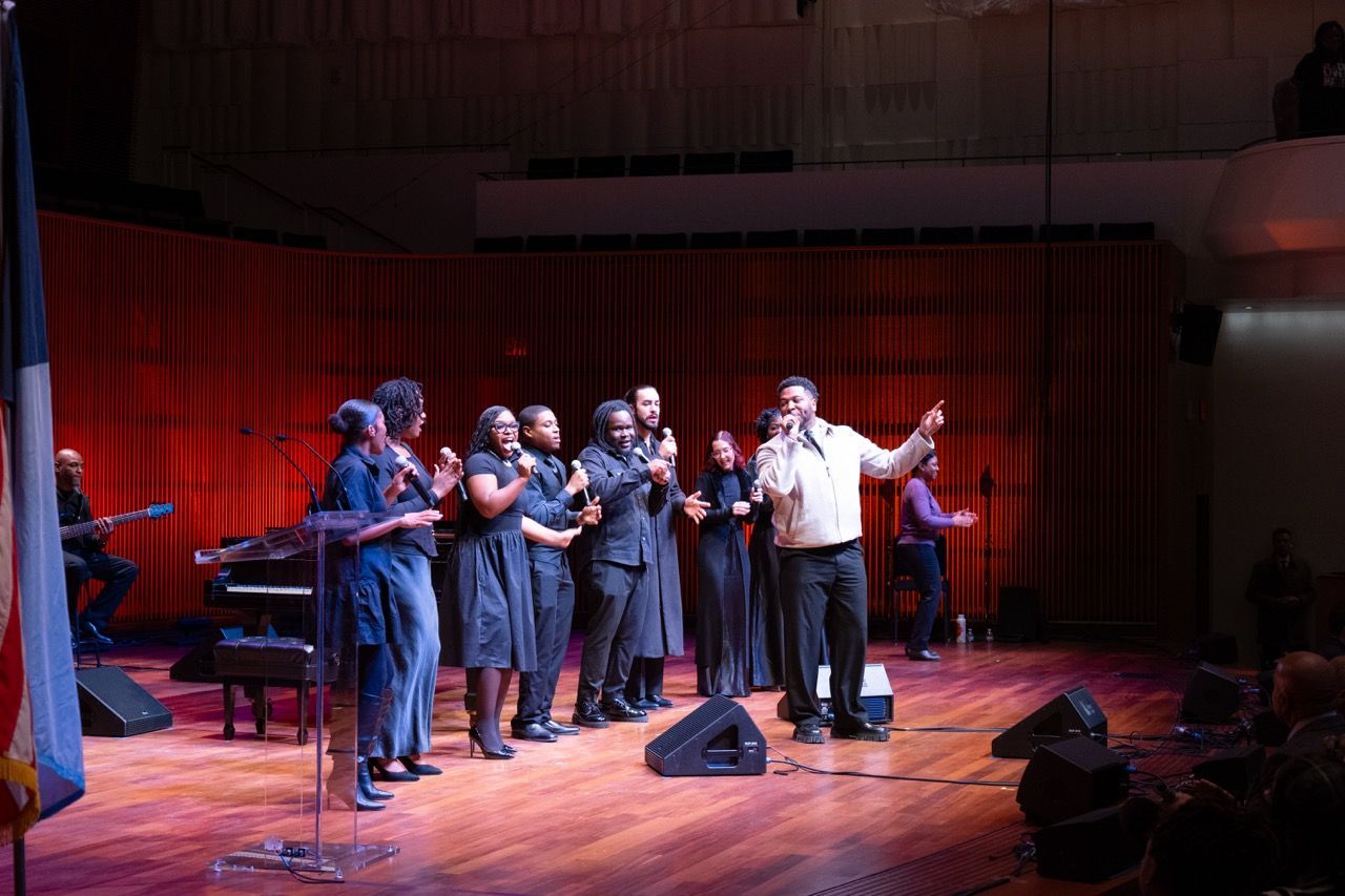 Group of eight singers standing on stage with microphones, performing with a guitarist seated in the background, in a concert hall with red lighting and wooden floors.